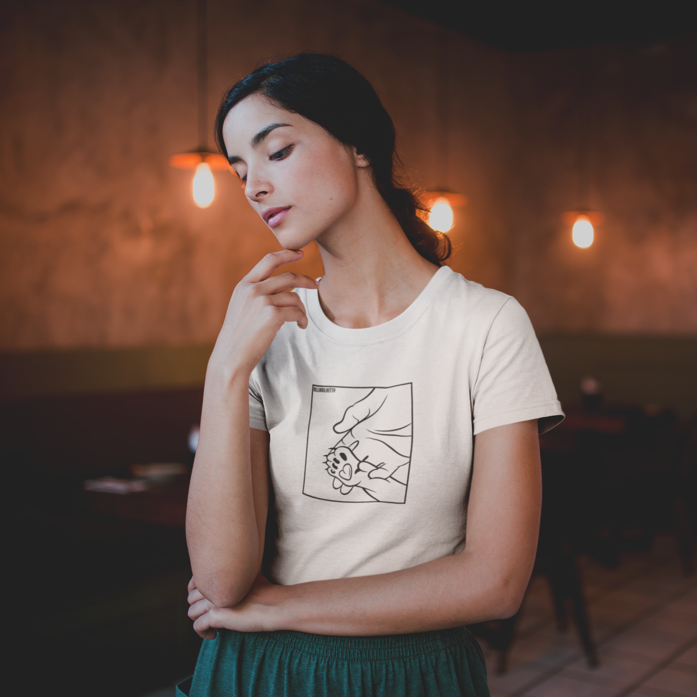 A woman wearing a white t-shirt with a graphic print of a paw and hand on the front, standing in a restaurant with dim lighting and a hood in the background.